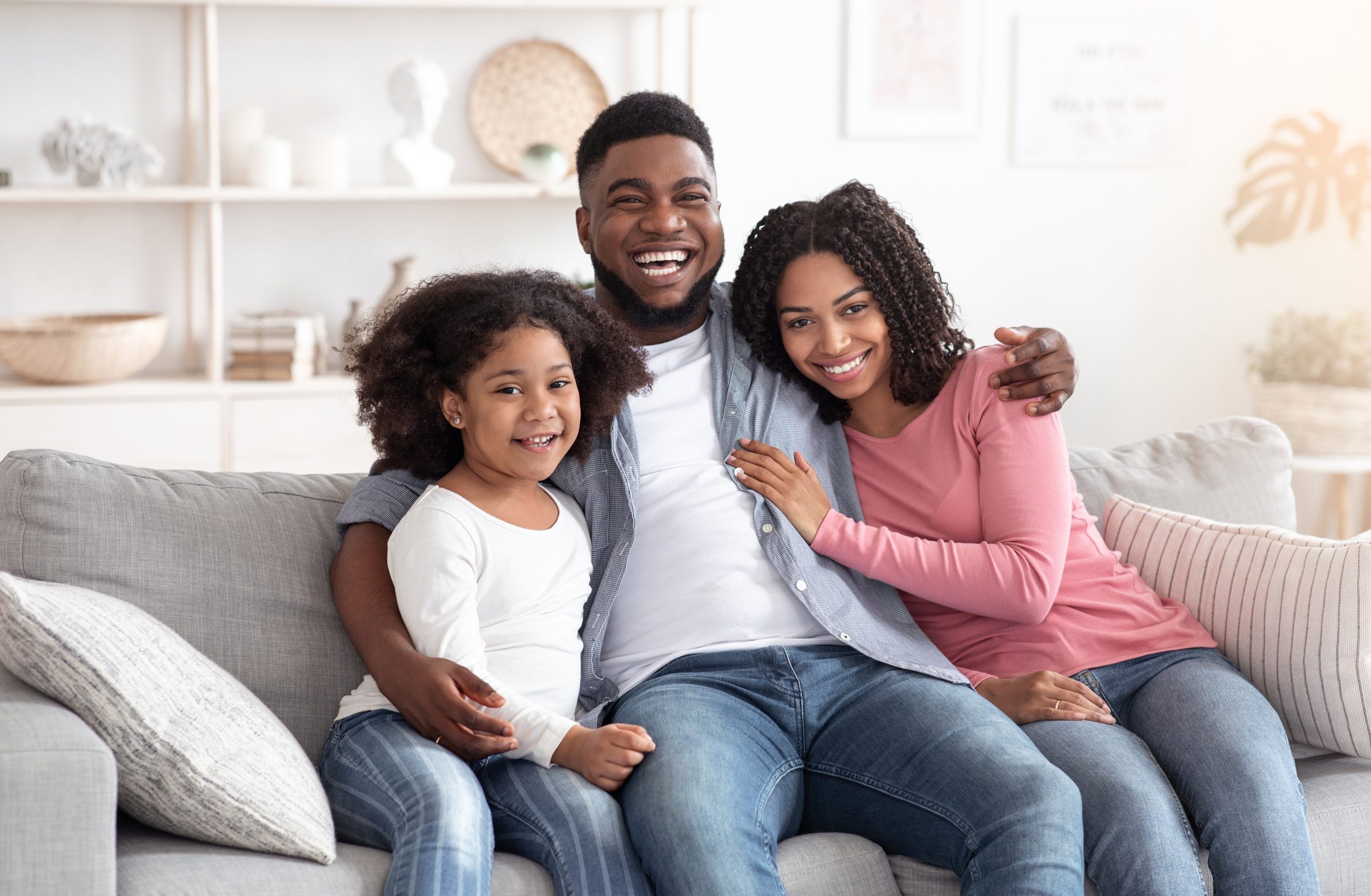 Family lifestyle portrait of happy black mom, dad with their little daughter at home, hugging on couch in living room and looking at camera